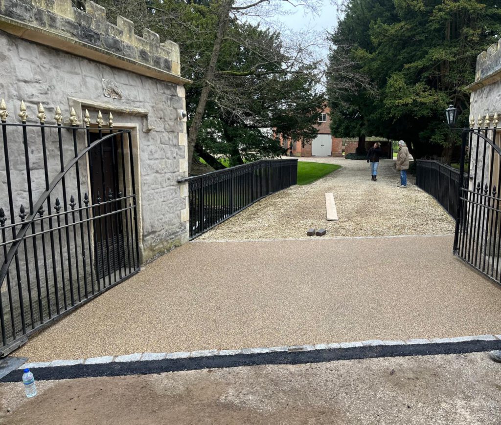 A worker in a high-visibility orange jacket inspects a newly laid gravel path, possibly the work of a paving contractor, nestled between stone walls with black metal gates. The path leads to a house among trees. A plank and bottle lie on the ground as another person walks away in the distance.