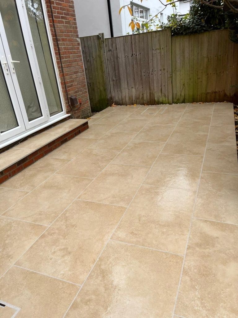 A small outdoor patio with large beige tiles, bordered by a wooden fence and brick walls, with steps leading to glass patio doors. The patio surface appears clean and slightly wet.