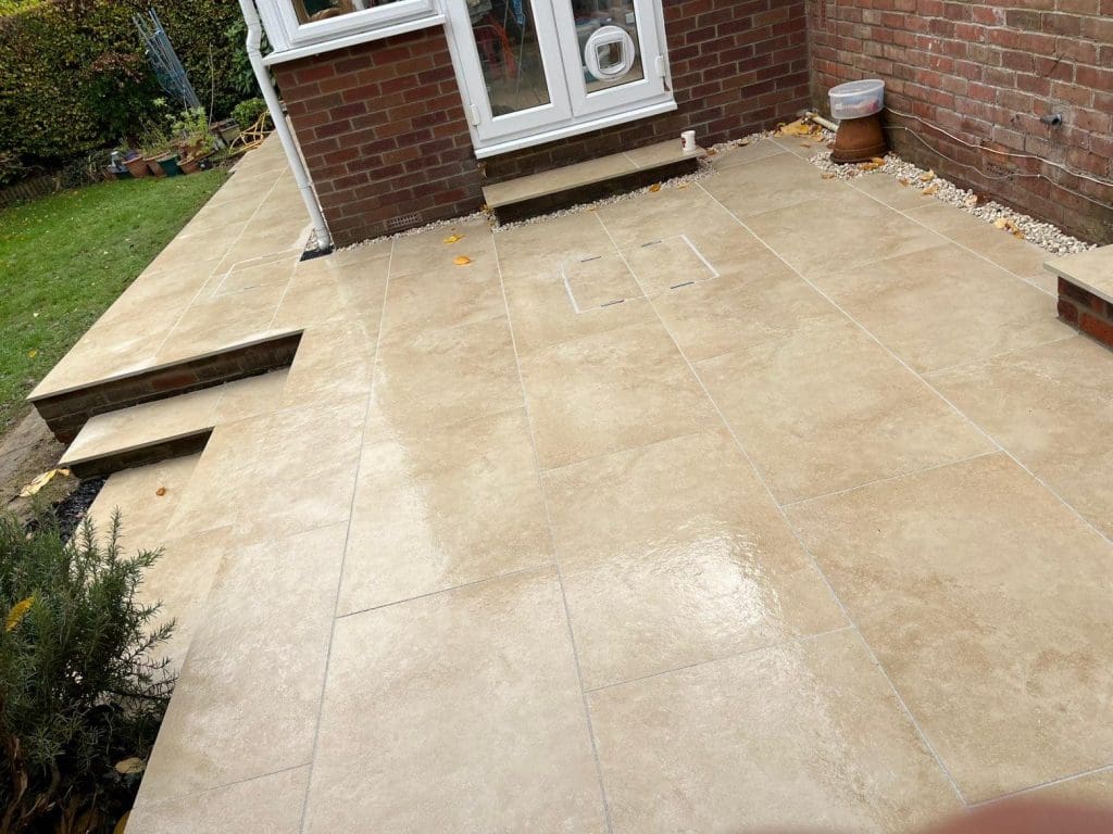 Large beige tiled patio with steps leading up to a brick house. A white door and window are visible. Some garden plants border the area, and there are small gravel sections along the edges.