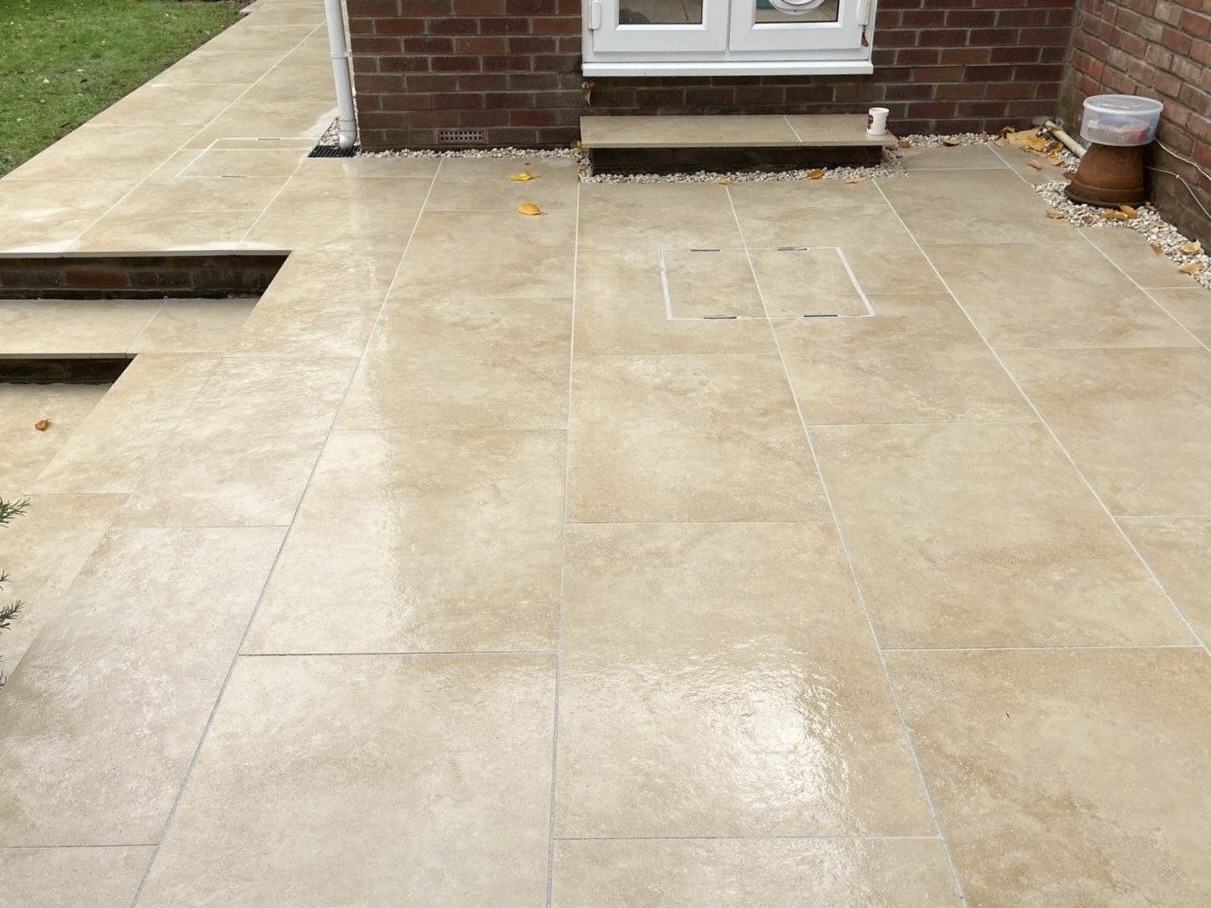 A beige-tiled patio with steps leading down to a lower area, bordered by a brick house. There are a few scattered leaves and a plant pot and light fixture near the wall.