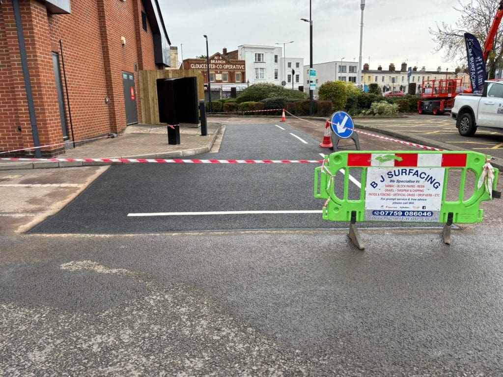 A section of freshly laid asphalt is blocked off with red and white tape and a green barrier labeled "BJ Surfacing." A construction area is visible in a parking lot beside a brick building.