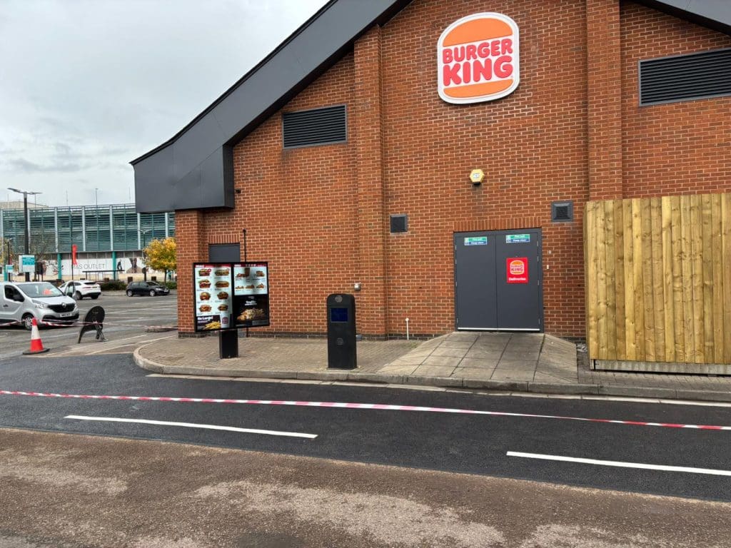 A Burger King drive-thru with a menu board and ordering speaker in front of a brick building. A red and white striped barrier tape blocks the drive-thru lane. A car and trees are visible in the background.