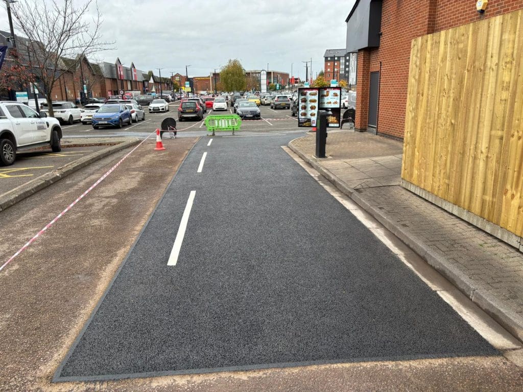 A freshly resurfaced section of road with new white lines runs between parked cars and buildings; a green barrier and traffic cones are positioned ahead, and traffic and pedestrians are visible in the background.