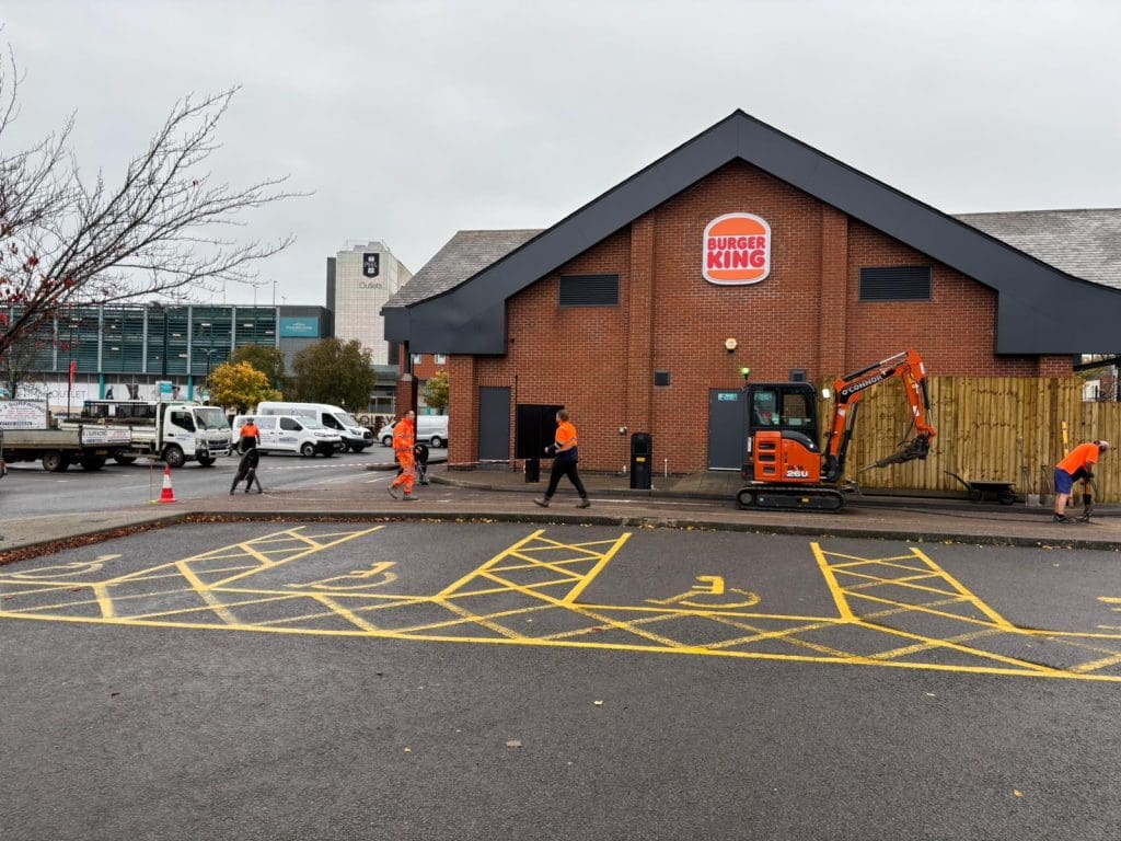 Several workers in orange vests work outside a Burger King; one operates a small excavator. The parking lot features yellow painted lines for disabled parking spaces. A tree and buildings are visible in the background.