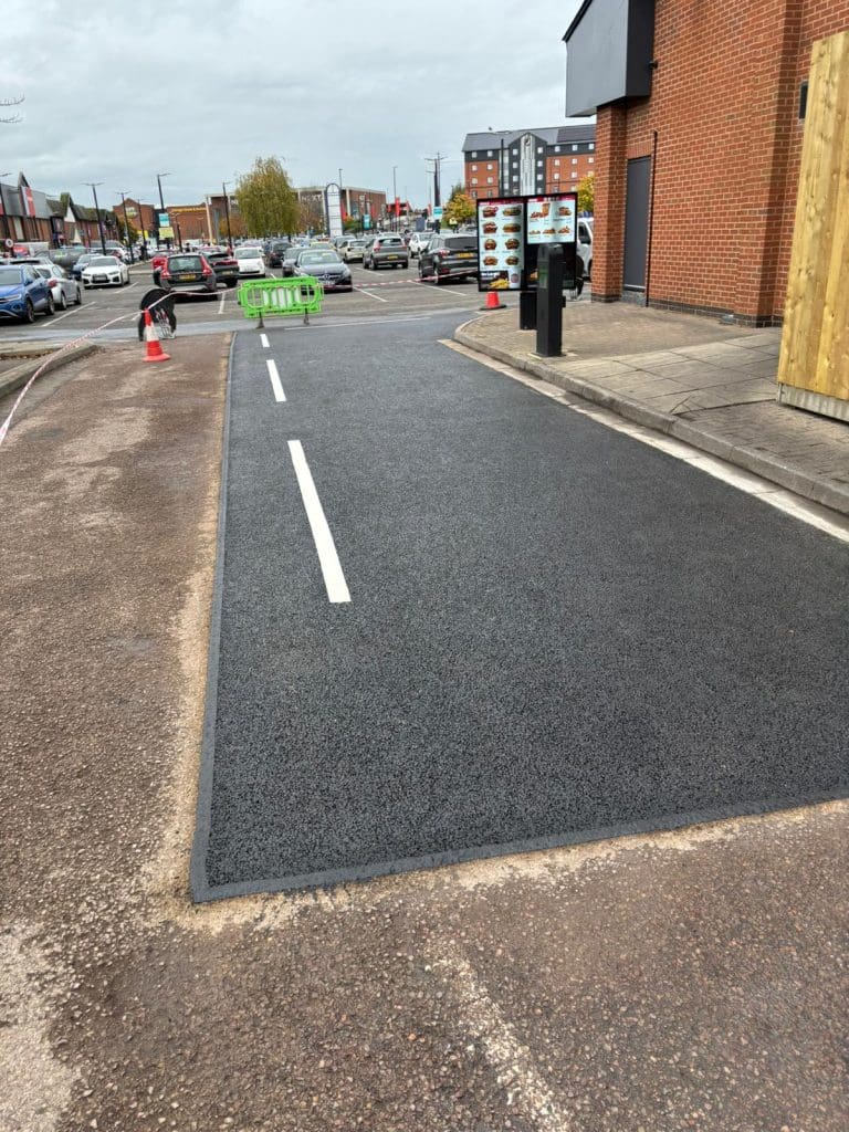 A newly paved drive-thru lane with fresh white road markings runs beside a red-brick building; traffic cones and barriers are visible, and cars are parked in the background under a cloudy sky.