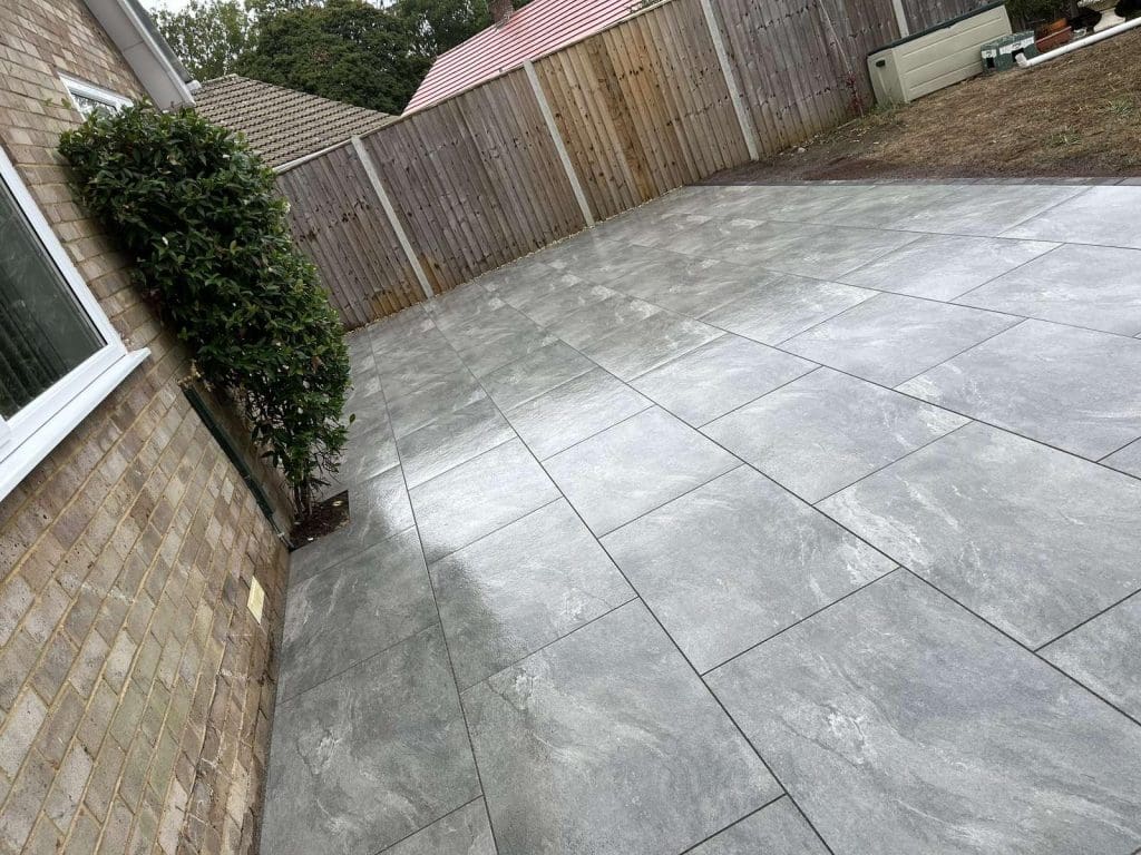 A backyard with large gray stone tiles forming a new patio beside a brick house, bordered by a wooden fence and a small leafy shrub near the house, with some bare soil in the corner.