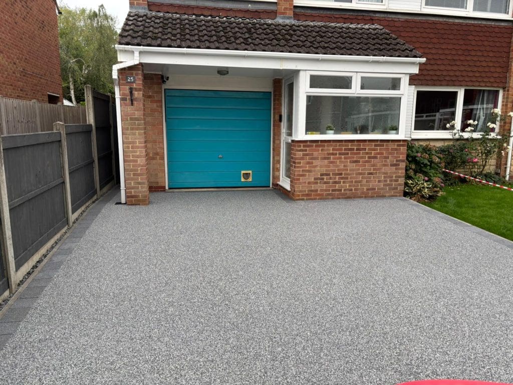 A house with a bright blue garage door, a small cat flap, and a newly laid light gray resin driveway. The front yard has green grass, flowers, and a black fence on the left side.