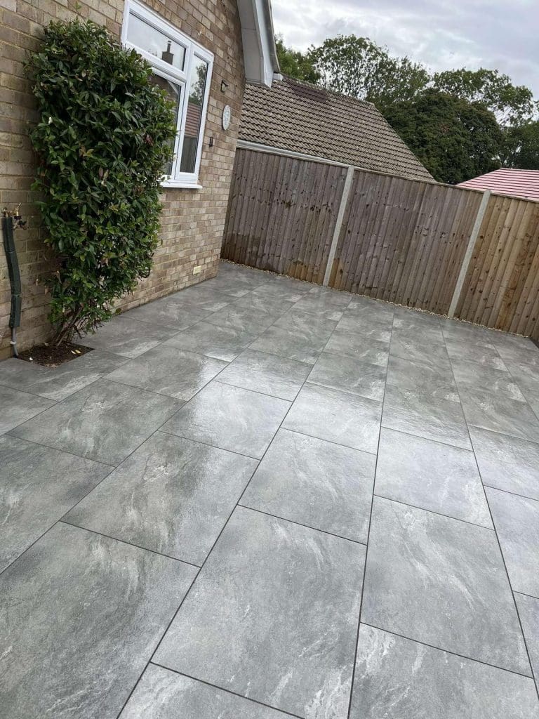 A clean patio with large gray stone tiles beside a brick house, bordered by a wooden fence and green hedge. Trees and neighboring rooftops are visible in the background.