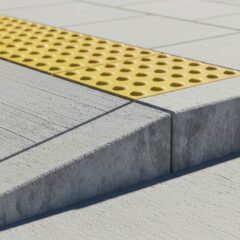 A concrete sidewalk curb ramp with a textured yellow tactile paving strip for visually impaired pedestrians. The paving has raised circular bumps.
