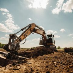 A large excavator digs into soil on a construction site under a blue sky with scattered clouds. Trees and green fields are visible in the background.