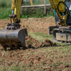 A small excavator with a yellow arm is digging soil in a grassy outdoor area. Only part of the machine and a person’s legs are visible.