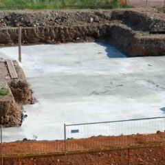A construction site with a freshly poured concrete foundation, surrounded by dirt, fencing, and some construction materials. Grass is visible in the background.