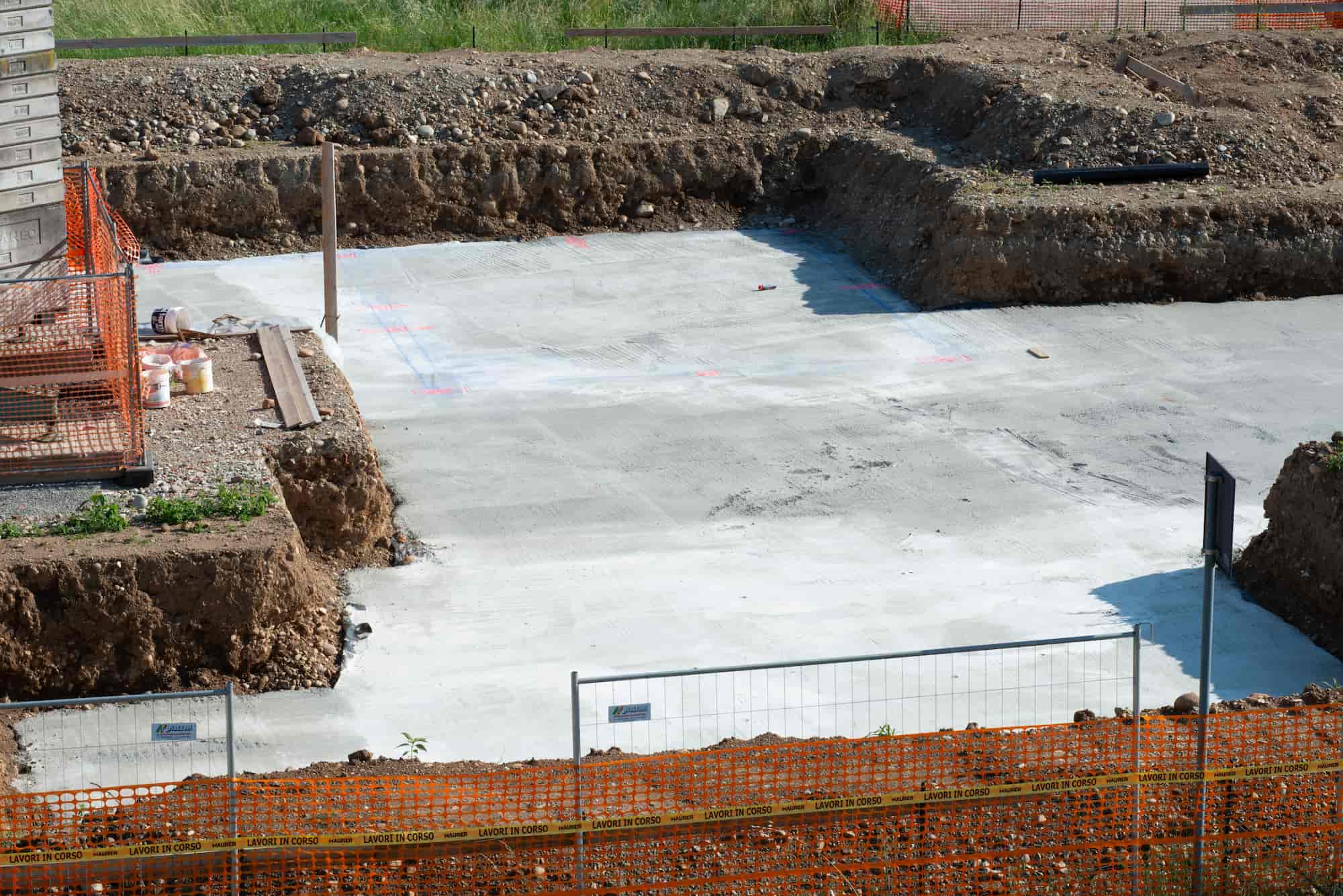 A construction site with a freshly poured concrete foundation, surrounded by dirt, fencing, and some construction materials. Grass is visible in the background.
