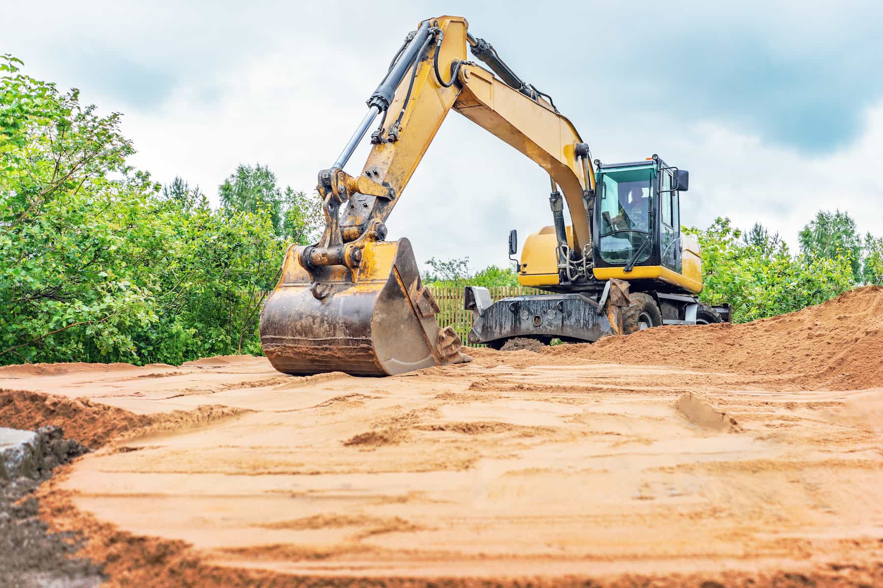 A yellow excavator moves a pile of dirt on a construction site surrounded by green trees under a cloudy sky.