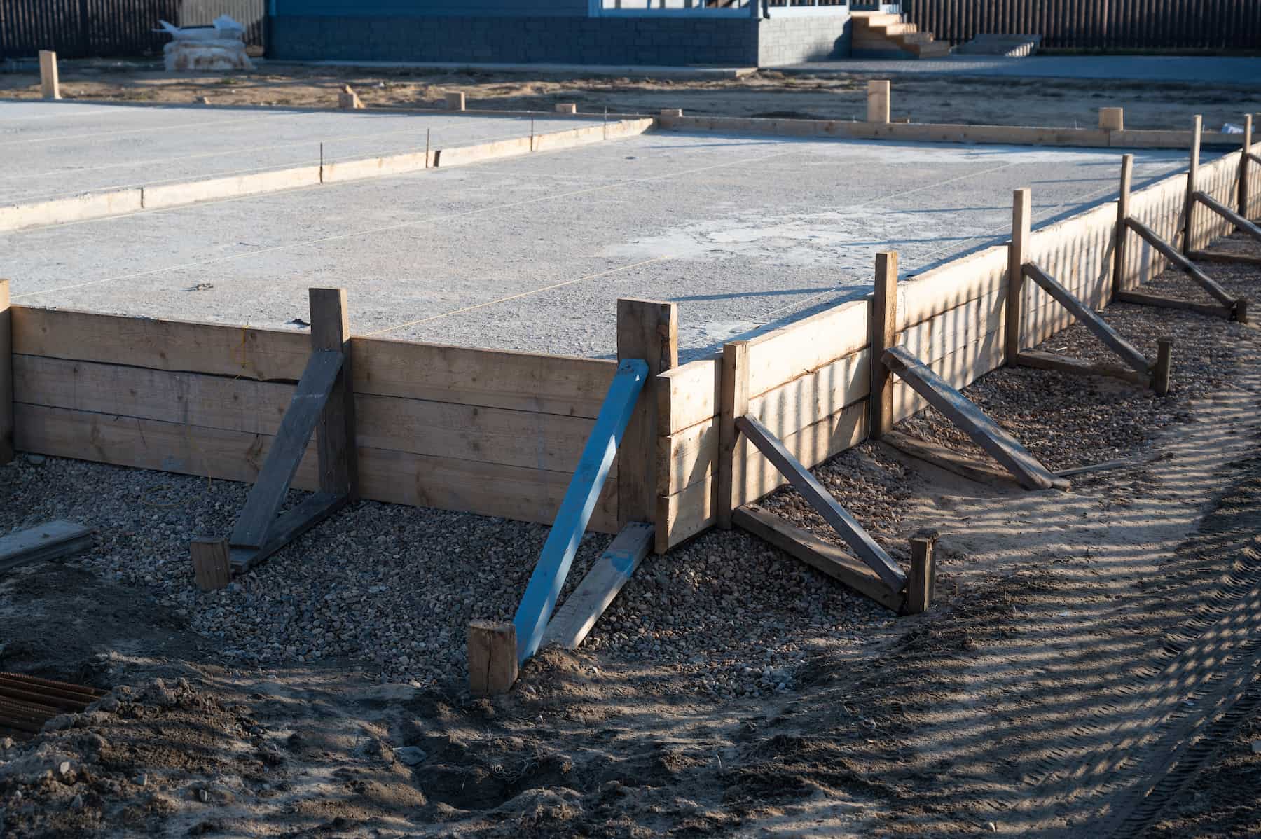 A concrete foundation slab bordered by wooden forms and supports at a construction site, with gravel and dirt surrounding the area.
