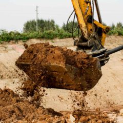 A yellow excavator dumps a load of brown soil onto the ground at a construction site with dirt and greenery in the background.