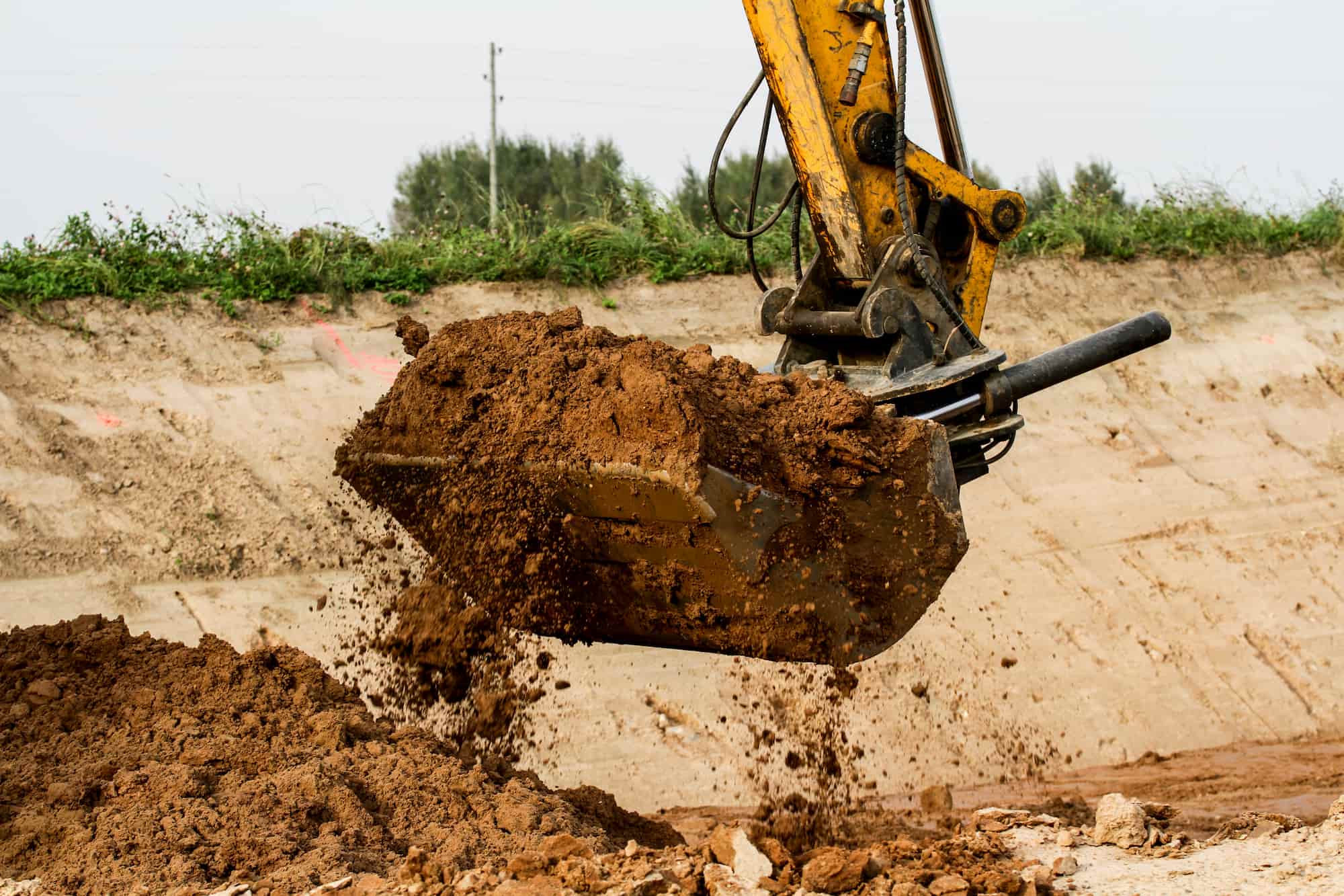 A yellow excavator dumps a load of brown soil onto the ground at a construction site with dirt and greenery in the background.