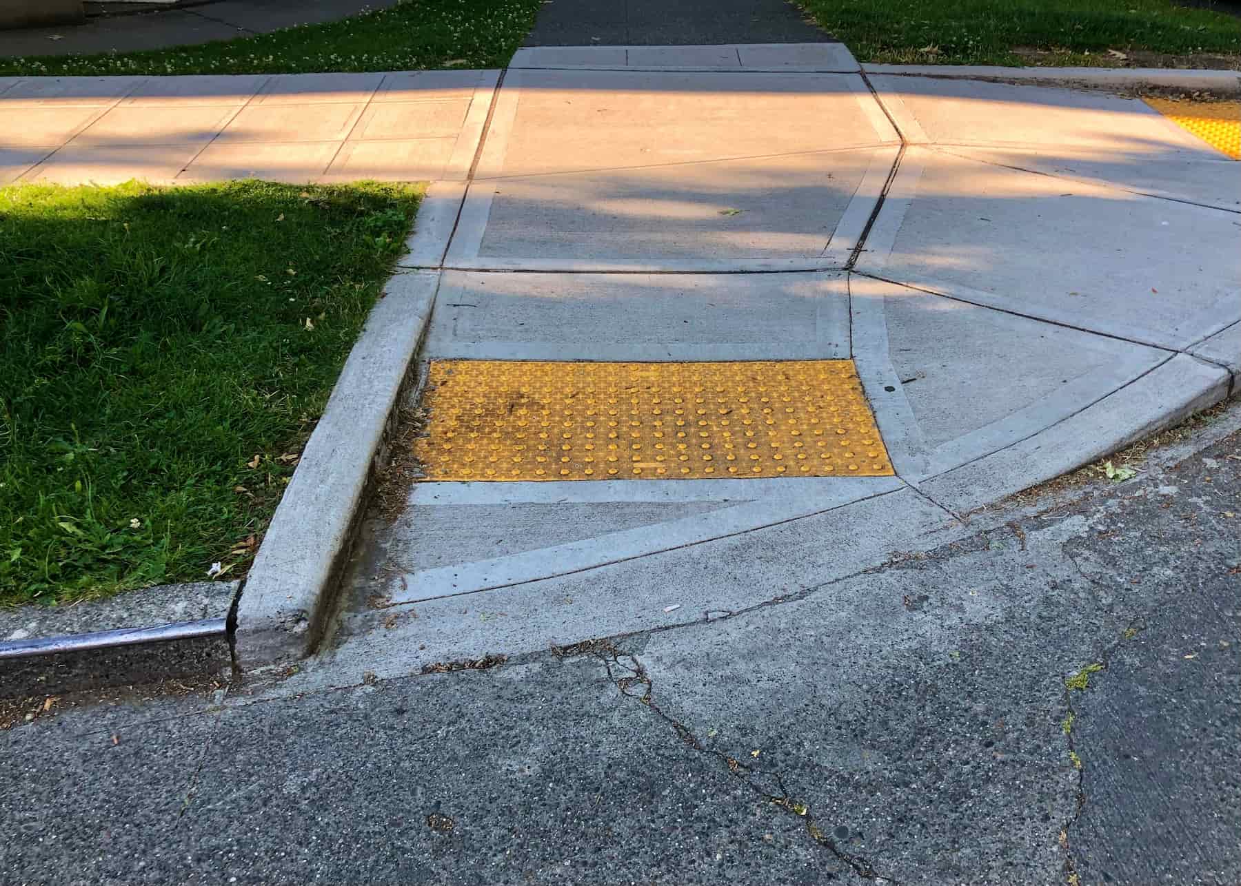 A sidewalk curb cut with a yellow textured tactile paving for accessibility, leading from the street to the sidewalk, bordered by concrete and grass.