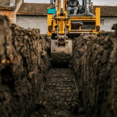 A yellow excavator digs a deep trench in muddy soil between two buildings, with the view focused on the bucket and the excavated earth.