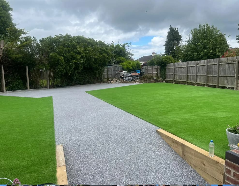 A backyard with artificial grass, a gravel or concrete path, wooden borders, a water bottle on a ledge, wooden fencing, and green trees under a cloudy sky.