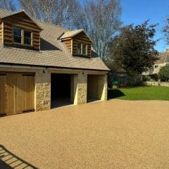 A detached garage with three wooden doors and dormer windows sits beside a grassy yard under a clear blue sky, bordered by trees and neighboring houses.