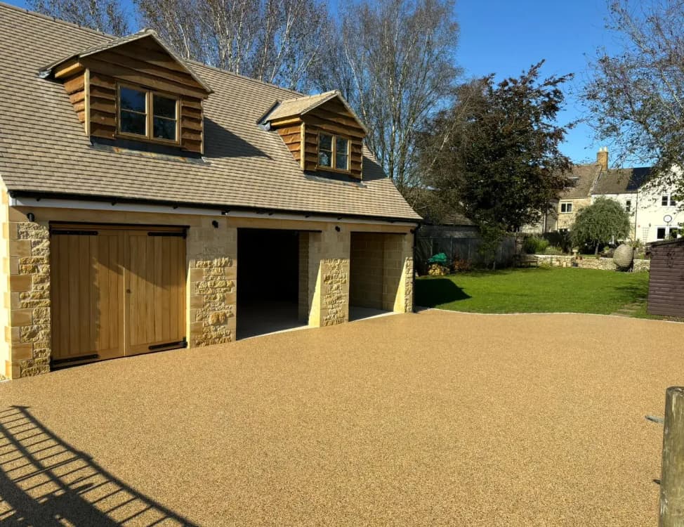 A detached garage with three wooden doors and dormer windows sits beside a grassy yard under a clear blue sky, bordered by trees and neighboring houses.