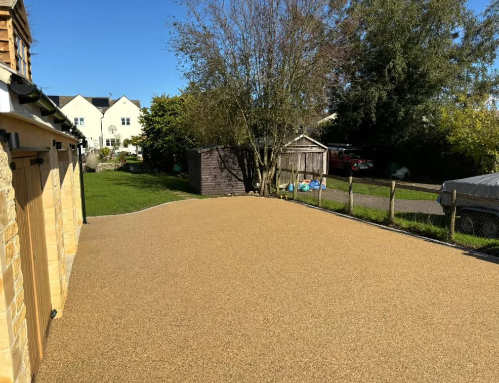 A tidy gravel driveway in front of a house, with a shed, trees, and a fenced yard in the background on a sunny day.