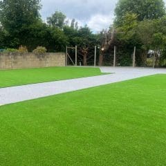 A yard with artificial grass sections, a strip of gray gravel or concrete path in the center, surrounded by trees and bushes under a cloudy sky.