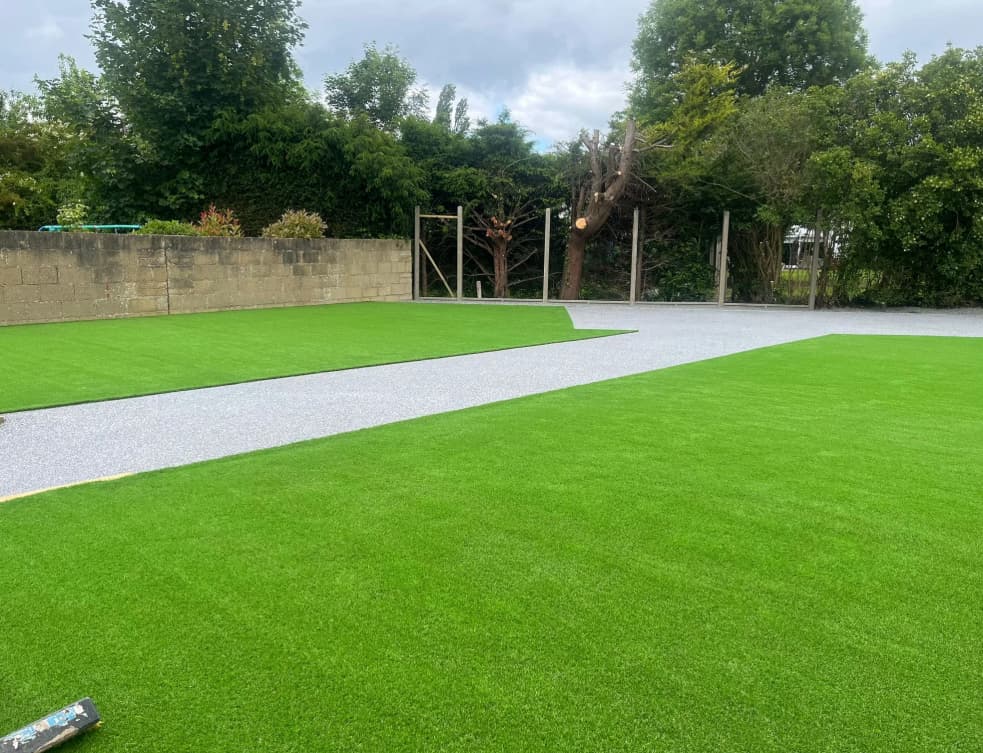 A yard with artificial grass sections, a strip of gray gravel or concrete path in the center, surrounded by trees and bushes under a cloudy sky.