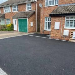 A newly surfaced black asphalt driveway in front of a brick house with green garage doors. A sign for "B J Surfacing" is on the right near the house. The sky is partly cloudy.