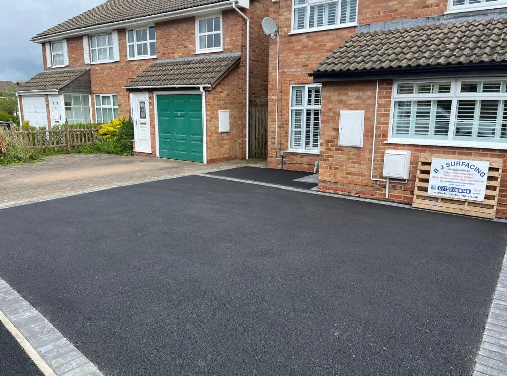 A newly surfaced black asphalt driveway in front of a brick house with green garage doors. A sign for "B J Surfacing" is on the right near the house. The sky is partly cloudy.