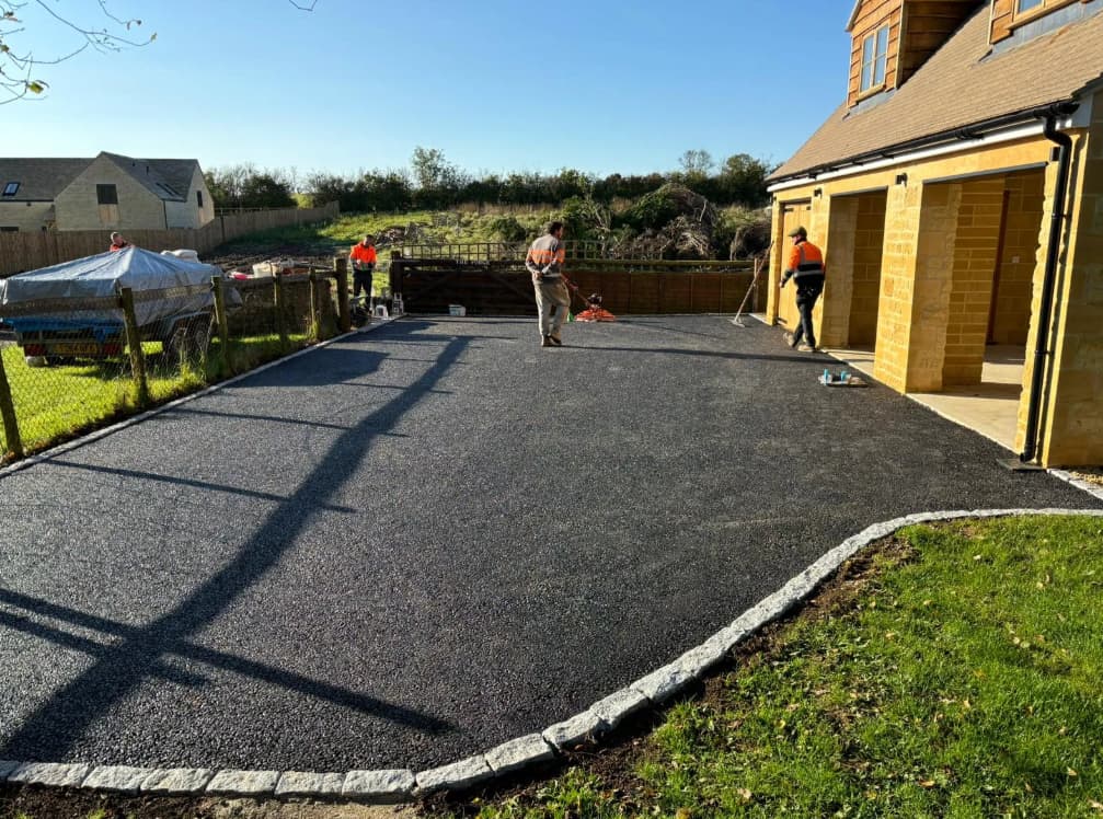 Three workers in orange vests spread asphalt on a driveway beside a house with a garage, while a grassy lawn and a wooden fence are visible nearby under a clear sky.
