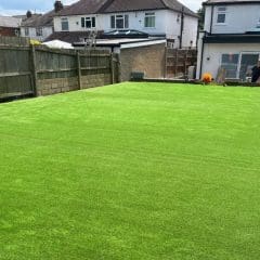 A backyard with bright green artificial grass, surrounded by a wooden fence and stone border, next to a white house with sliding glass doors.