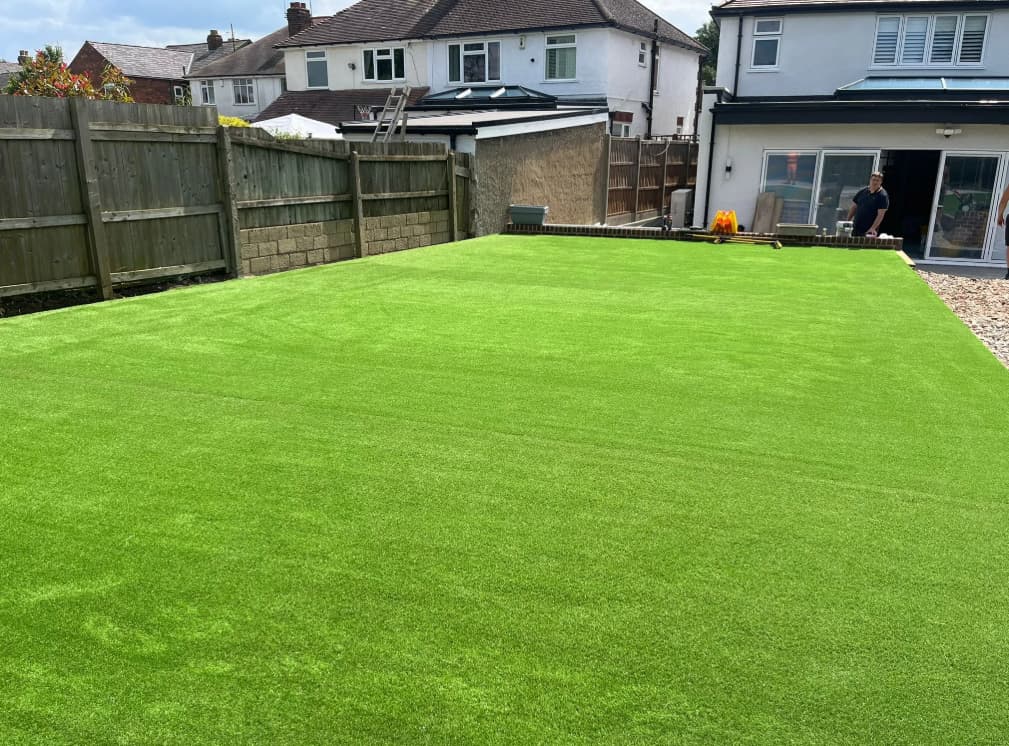 A backyard with bright green artificial grass, surrounded by a wooden fence and stone border, next to a white house with sliding glass doors.