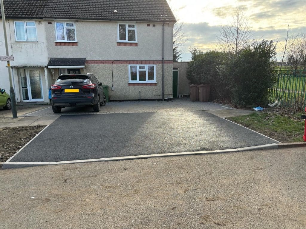 A freshly paved driveway leads to a parked black car beside a beige house with white windows and brown trim. There are brown bins and bushes to the right and a green lawn on the left.