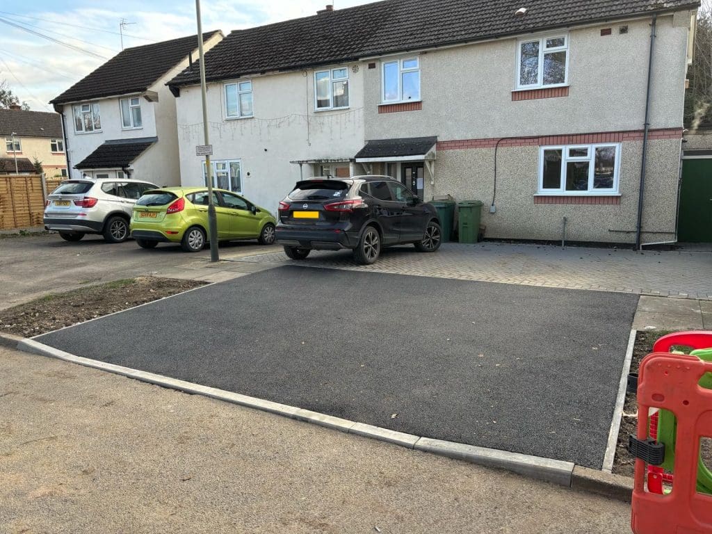 A freshly paved asphalt driveway is in front of a house with two parked cars. There are bins near the house, and a red and green barrier is seen at the edge of the image.