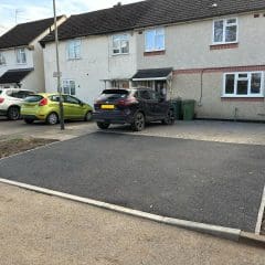 A freshly paved asphalt driveway is in front of a house with two parked cars. There are bins near the house, and a red and green barrier is seen at the edge of the image.